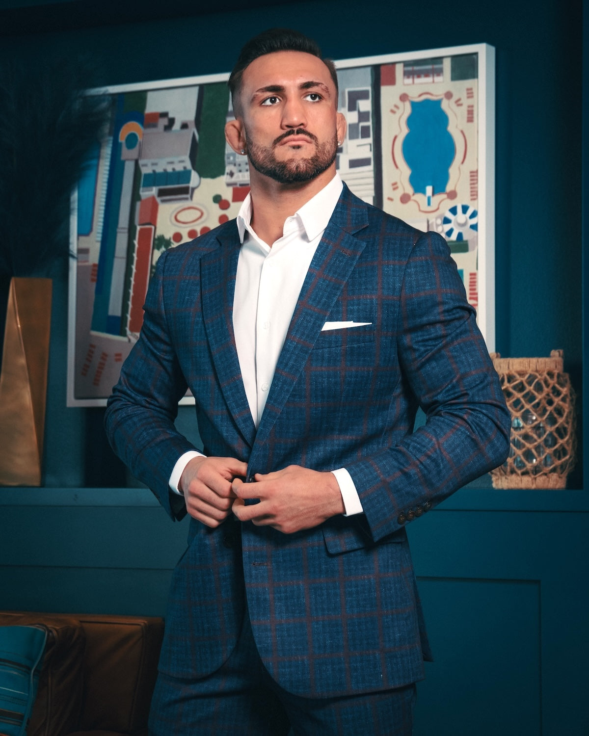Man wearing a blue plaid suit with a white shirt in a room with decorative wall art.