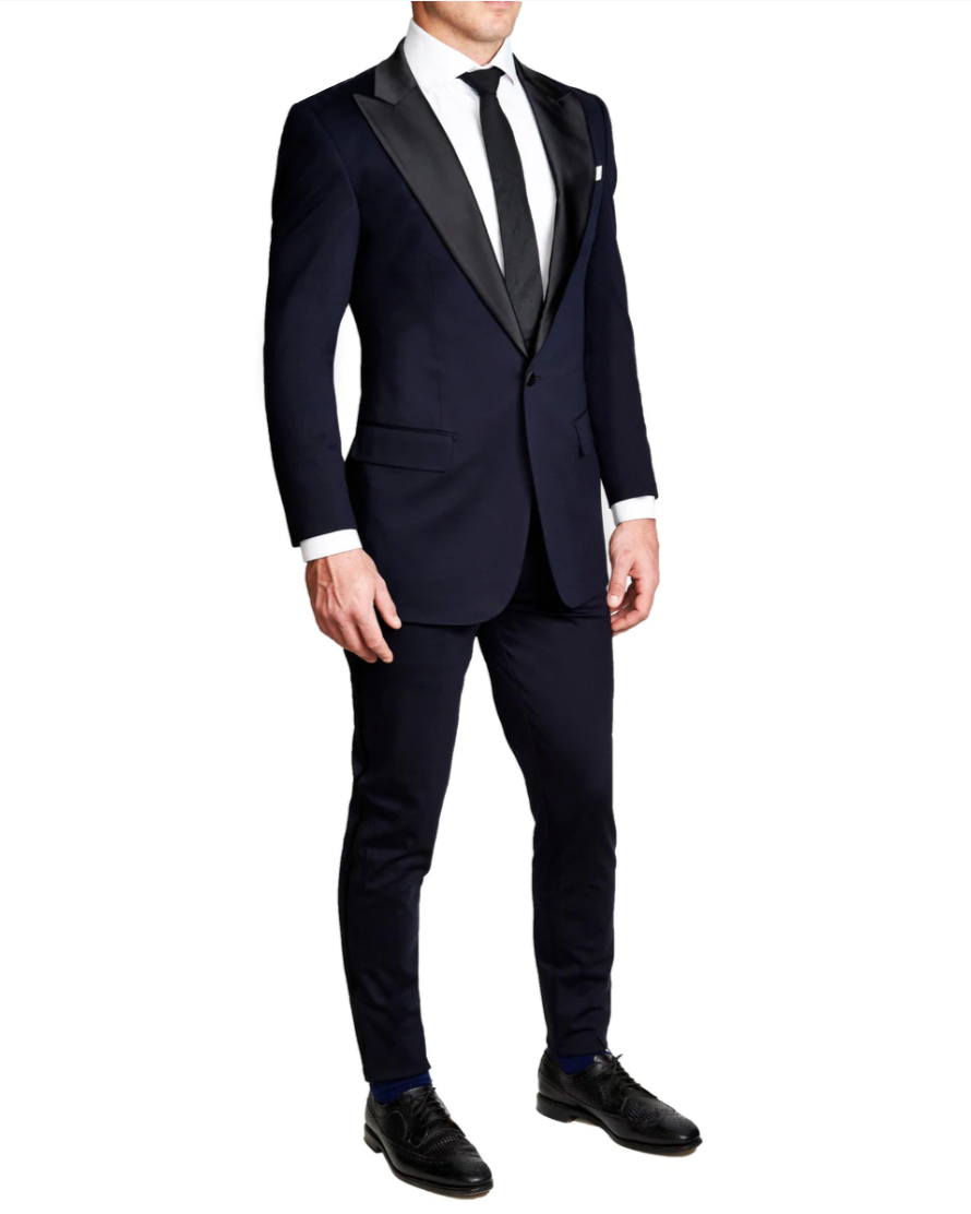 A man wearing a navy tuxedo with peak lapels, consisting of a jacket and trousers, standing against a white background.
