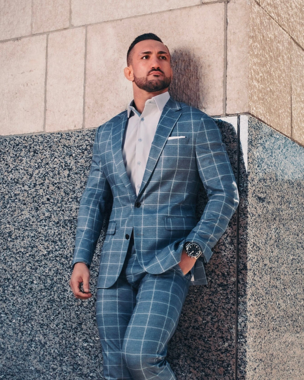Man in a blue checkered suit standing against a building wall.
