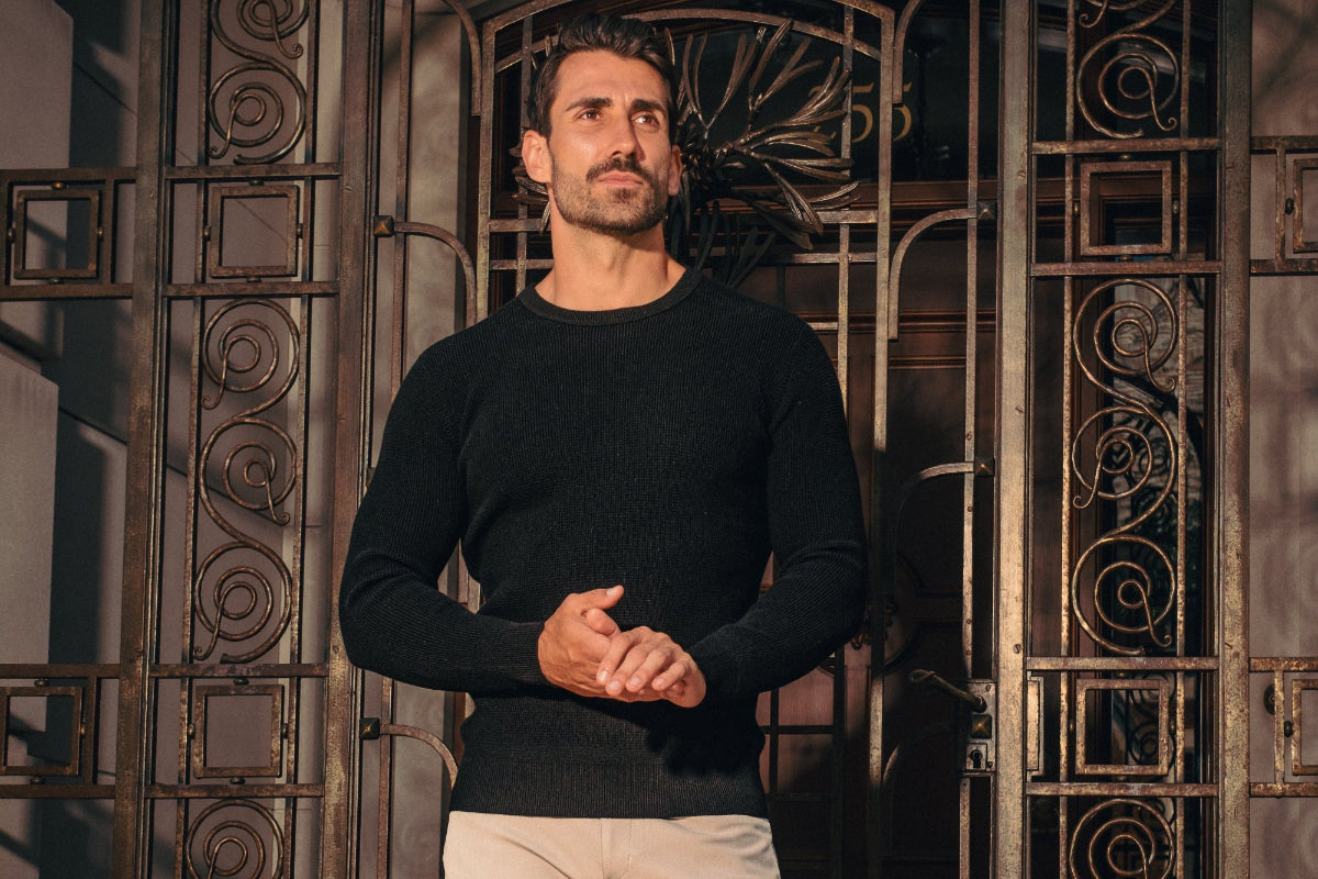 Man wearing a black sweater and light-colored pants standing in front of an ornate metal gate.