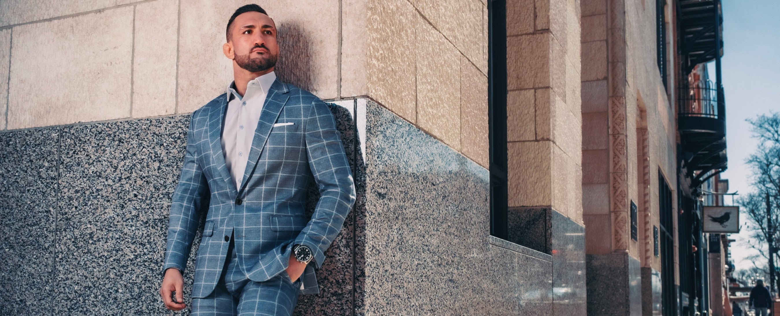 Man in a blue checkered suit standing against a building wall.